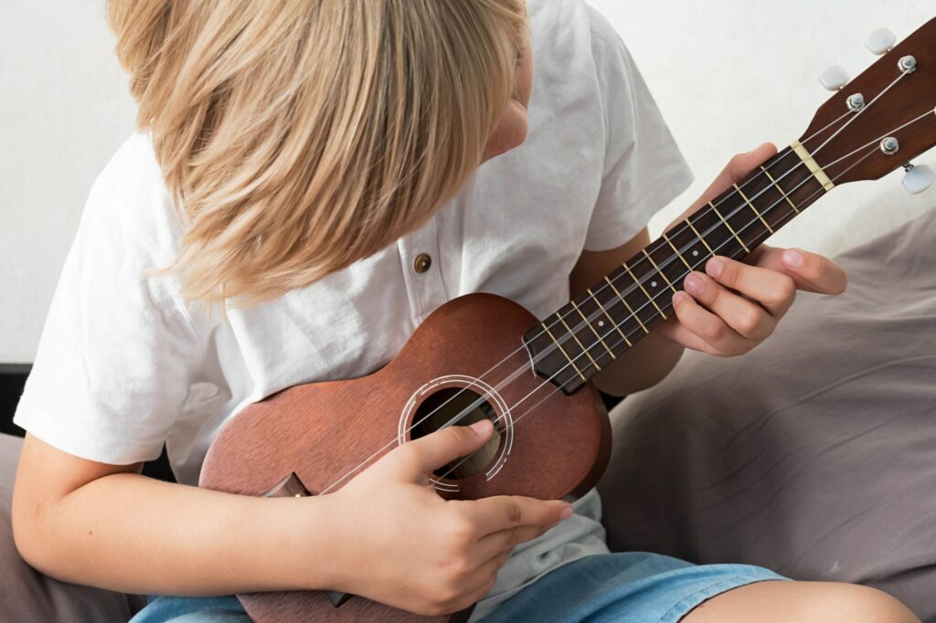 Young Boy Tuning Ukulele at Home. Boy Sitting on Couch Playing Acoustic Guitar.