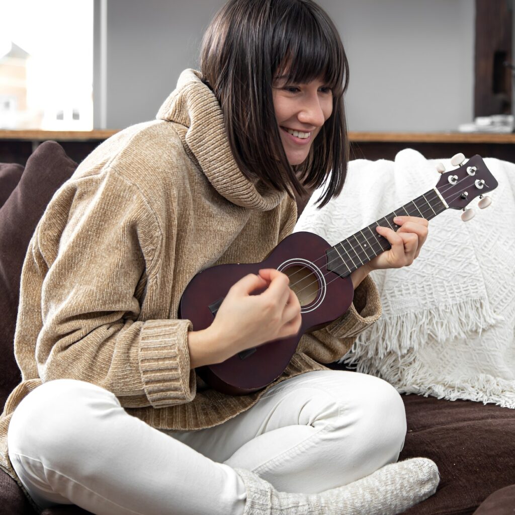 Young beautiful woman learning to play ukulele at home with online lessons.